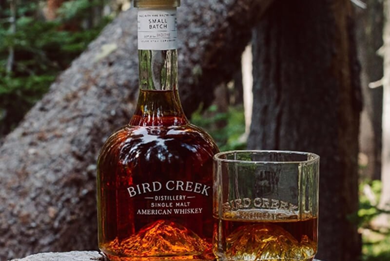 Bird Creek single malt whiskey bottle and glass on a rock in a forest