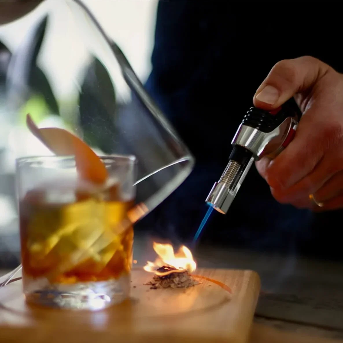 Torch lighting wood chips under a glass cloche beside an old fashioned cocktail